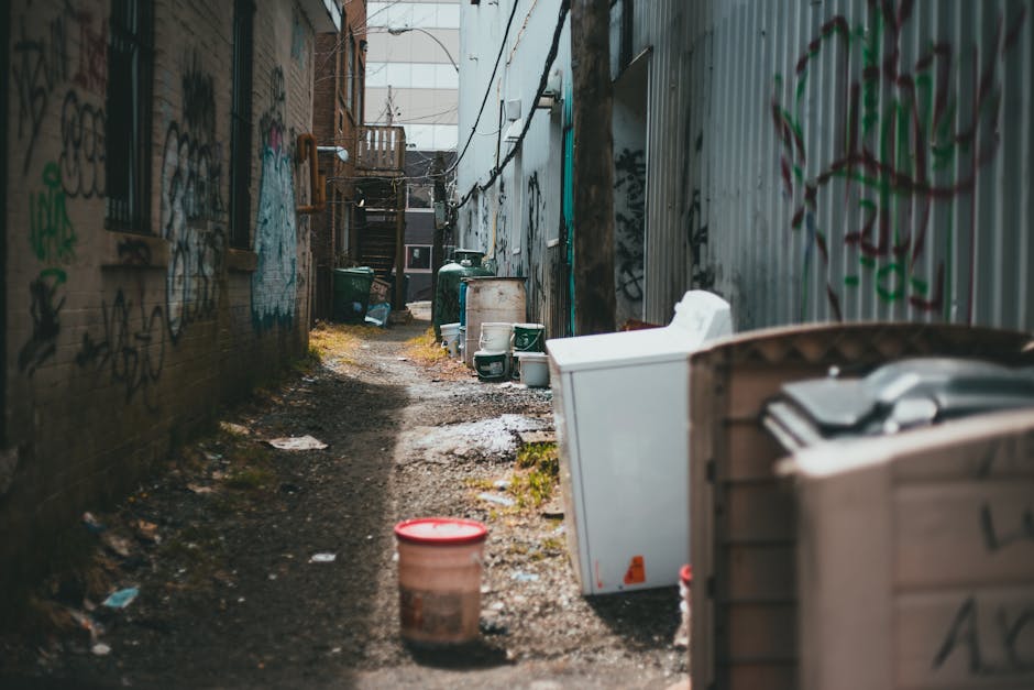 A narrow alleyway between two brick and metal-sheet buildings is cluttered with discarded household items and waste. On the right side, there is a white washing machine and a partially visible cardboa