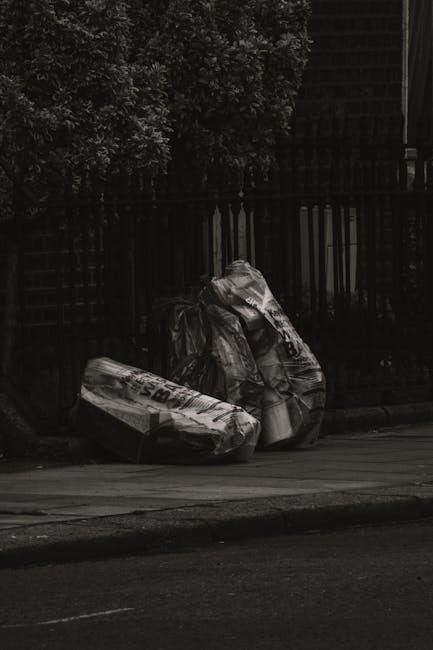 A narrow alleyway between two brick and metal-sheet buildings is cluttered with discarded household items and waste. On the right side, there is a white washing machine and a partially visible cardboa
