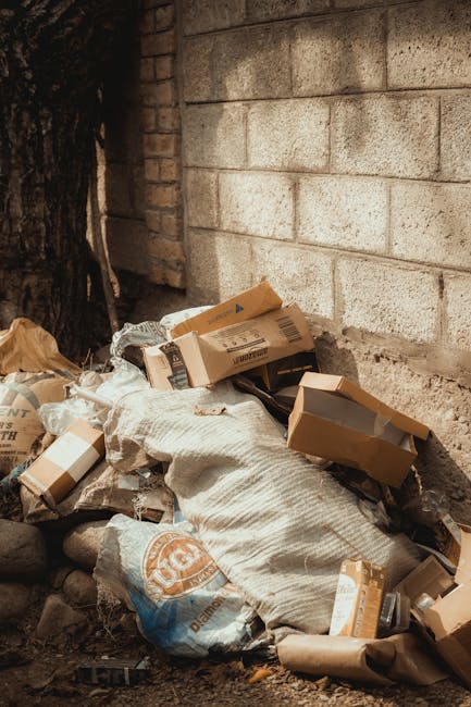 A pile of waste materials is accumulated at the base of a brick wall in an outdoor setting, consisting mainly of flattened cardboard boxes, some with printed labels and barcodes, mixed with white wove