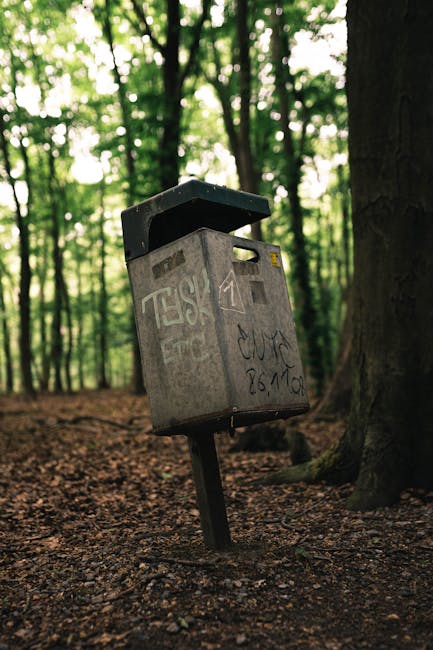 A tall, rigid plastic blue rubbish bin with a closed lid, situated outdoors on a patch of grass and accompanied by another waste container visible on the left, which is primarily red with black polka